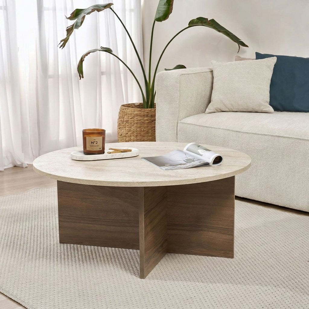 Round coffee table with travertine top and dark wooden base in cross shape. Bench and plant in the background.
