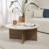 Round coffee table with travertine top and dark wooden base in cross shape. Bench and plant in the background.